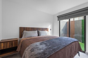A modern bedroom with a wooden headboard, a large bed with a patterned blanket, a nightstand, and a sliding glass door leading to a garden. - Modern Apartment with Pool in Querétaro (El Marques)