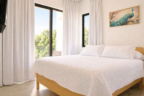 Une chambre à coucher avec un grand lit, des rideaux blancs, un tableau représentant un paon et une vue sur la verdure à l’extérieur.