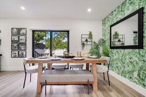 Un coin repas avec une table en bois dressée pour quatre personnes, des chaises à revêtement gris et un banc. Les murs sont ornés de tableaux encadrés, il y a un grand miroir et un papier peint vert à motifs de feuilles.