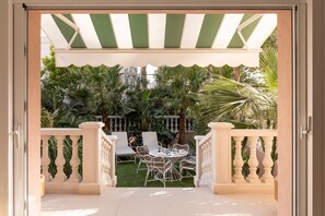 Eine Terrasse im Freien mit einem weiß-grün gestreiften Markisenvorzelt, einem runden Tisch mit Stühlen und einem üppigen Garten mit verschiedenen Pflanzen.