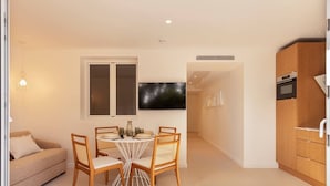A modern living room with a round dining table, wooden chairs, a sofa, a TV, and a kitchen area with cabinets and a microwave. - Carla Rosa 5 Apartement Terrazza - 2 BDR Piscine (Cannes)