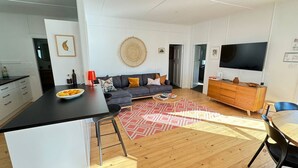 A modern living space with a kitchen, dining area, and a large TV. - Bendy Beach House, beach side property at Washerwomans beach and Sting Ray Bay (Bendalong)