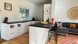 A modern kitchen with white cabinetry, a black countertop, and a central island. There is a dining area with a gray sofa and a red lamp. - Bendy Beach House, beach side property at Washerwomans beach and Sting Ray Bay (Bendalong)
