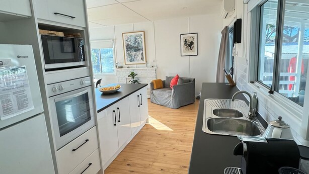 A modern kitchen with white cabinetry, a black countertop, and a built-in oven. There is a dining area with a gray sofa and a small table. - Bendy Beach House, beach side property at Washerwomans beach and Sting Ray Bay (Bendalong)