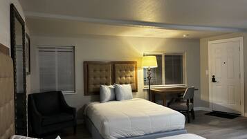 A hotel room with a bed, a desk, a chair, a lamp, and a door.