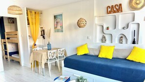 Et moderne, lyst værelse med køjeseng, spiseplads med bord og stole, et rundt bord med en plante og en sofa med gule puder.
