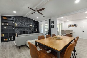 A modern living room with a fireplace, a dining area with a wooden table and chairs, and a built-in bookshelf. - Spacious Updated 4-Bed Home in Irving (Irving)