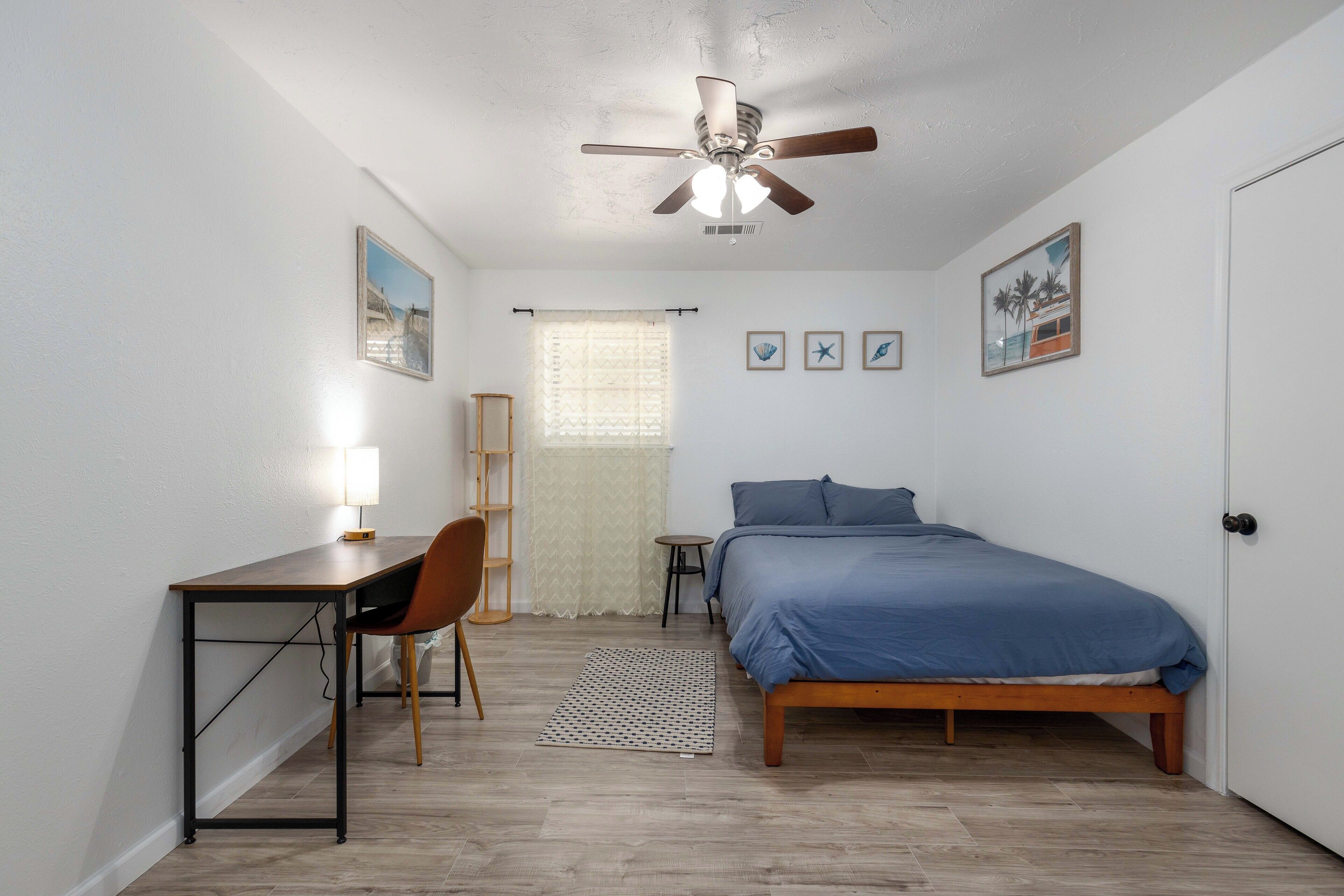 A bedroom with a bed, desk, chair, ceiling fan, and a window with curtains.