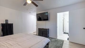 Une chambre à coucher comprenant un lit, un ventilateur de plafond, un téléviseur fixé au mur, une commode et une porte donnant sur une salle de bain.