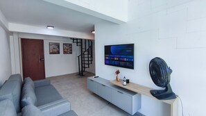 A modern living room with a grey sofa, a wooden TV stand, a mounted TV, a standing fan, and a staircase leading to another level. - Max Carneiros Suítes  (Tamandaré)