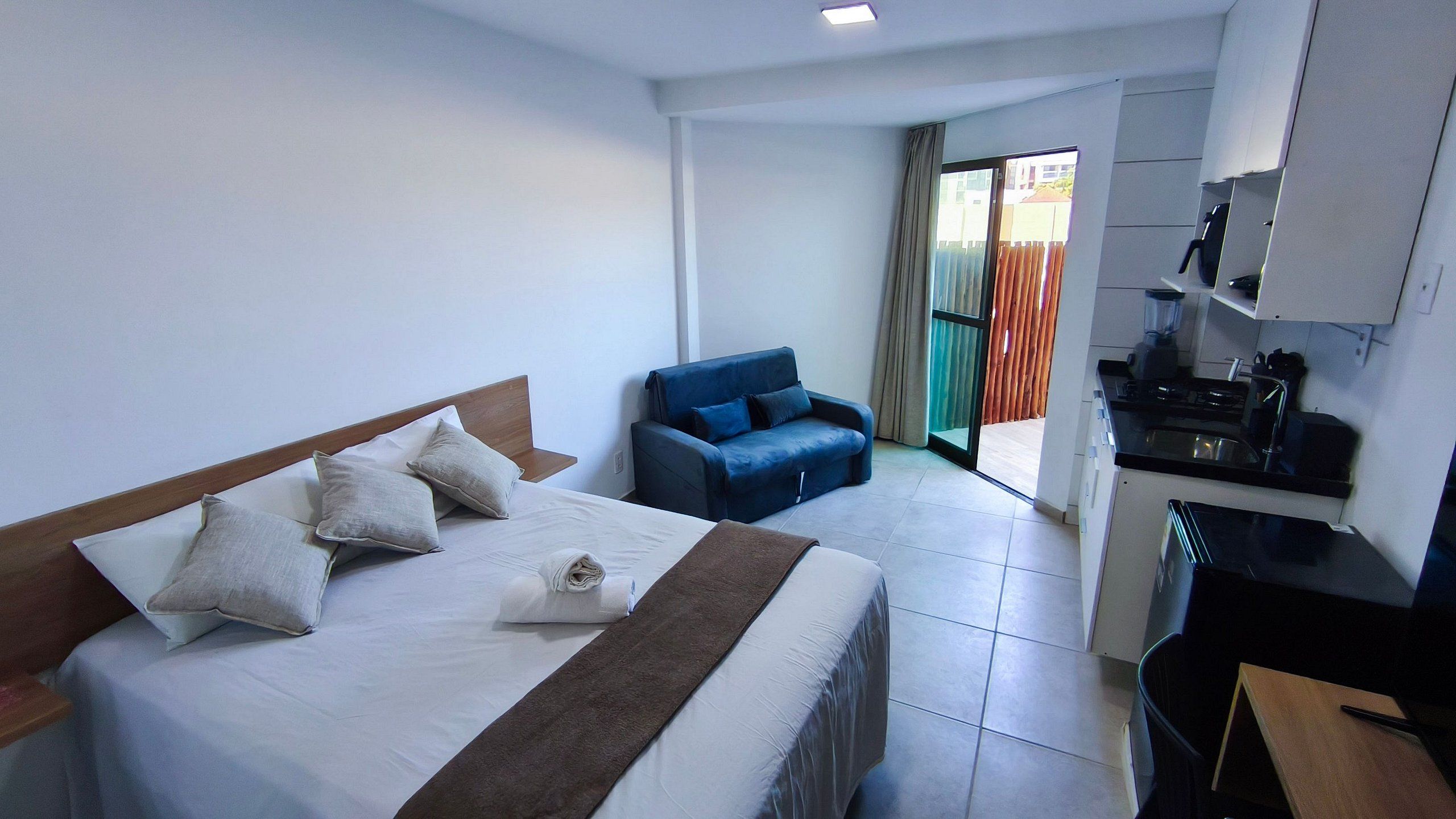 A hotel room with a bed, a blue armchair, a kitchenette, and a sliding glass door leading to an outdoor area.