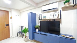 Ein Zimmer mit Fernseher auf einem blauen Schrank, einer Topfpflanze, einem kleinen Hocker und einer Tür.