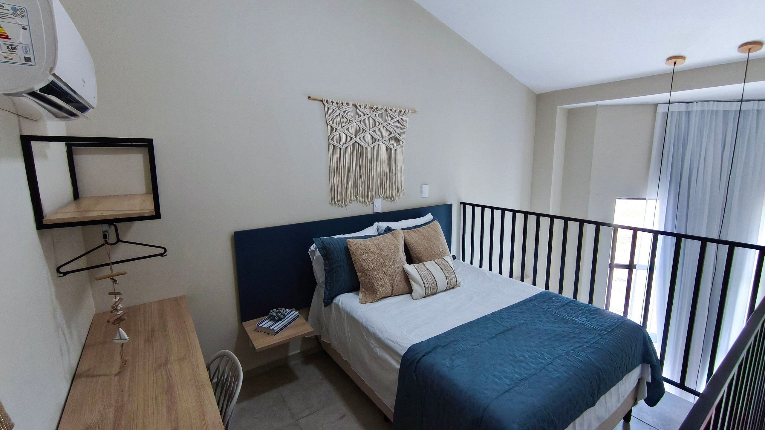 Ein Schlafzimmer mit Bett, Schreibtisch, Stuhl, einer Wanddekoration aus Makramee und einem Balkon mit Vorhängen.