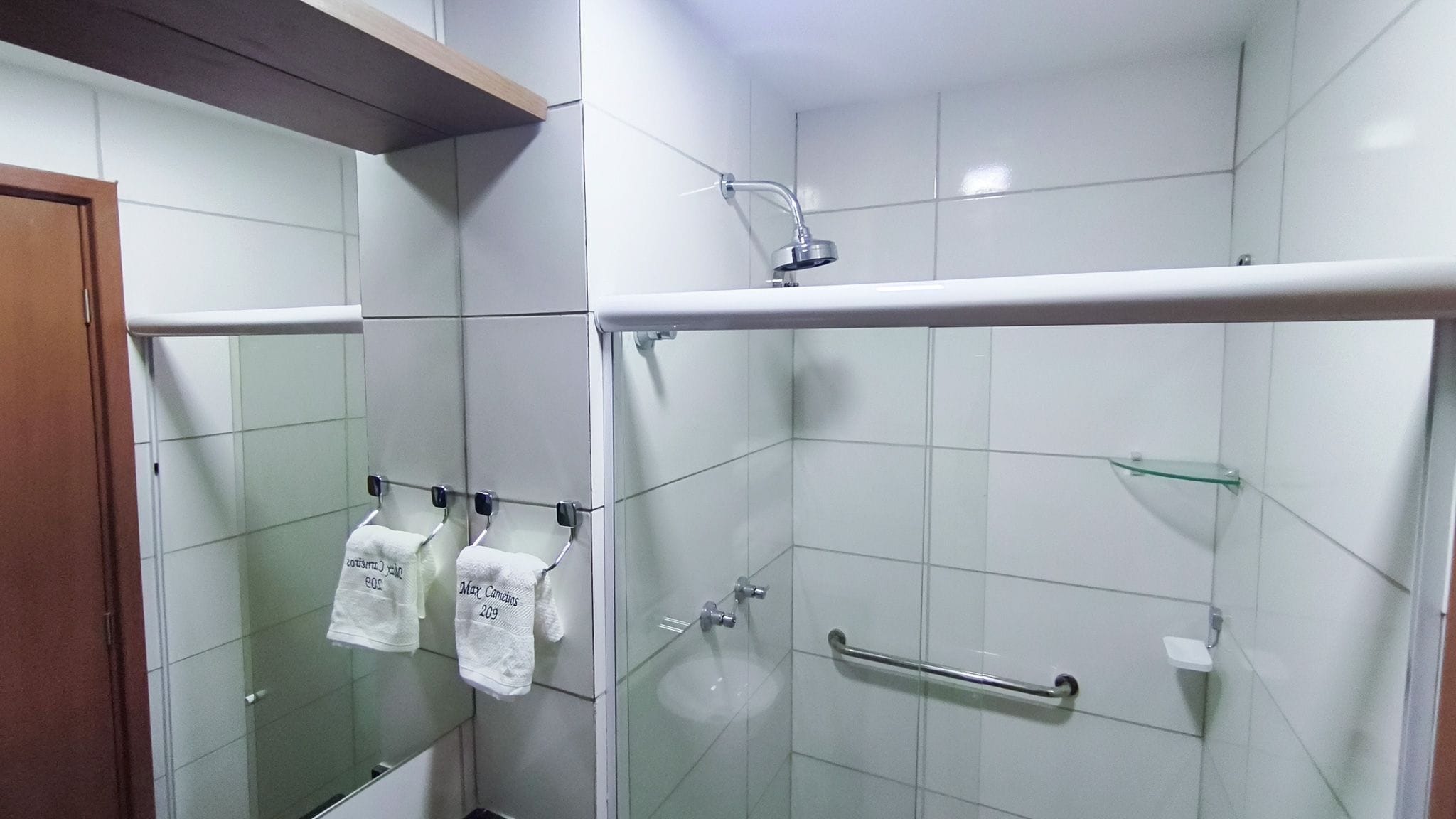 Ein Badezimmer mit weißen Fliesen, einer Duschkabine aus Glas, einem Waschbecken aus Glas und zwei Handtüchern, die an Haken hängen.