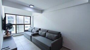 A modern living room with a grey sectional sofa, a flat-screen TV mounted on the wall, and a large window providing natural light. - Max Carneiros Suítes  (Tamandaré)