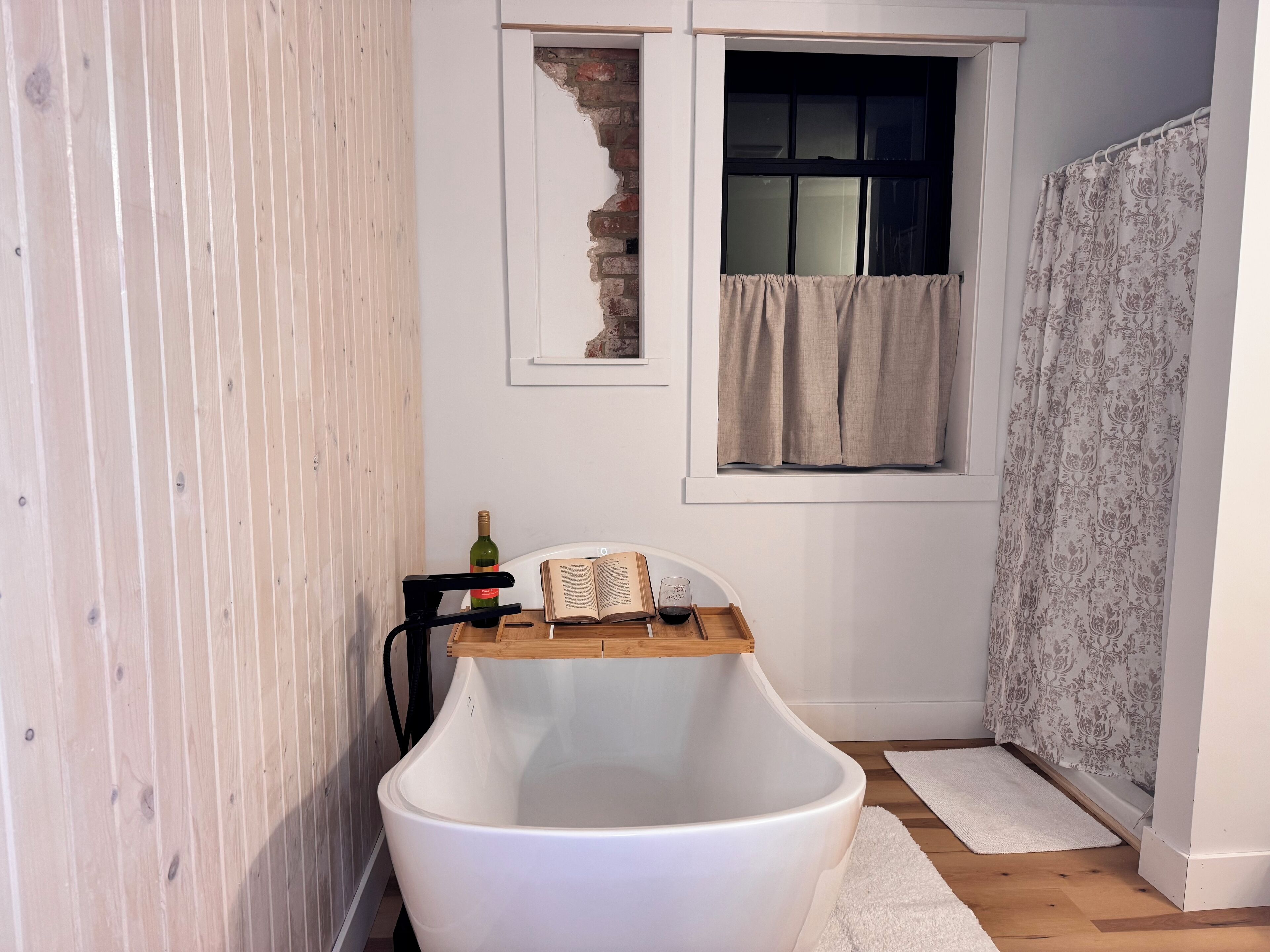 A bathroom with a freestanding bathtub, a window with curtains, a wooden floor, and a white wall.