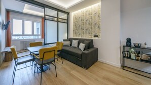 Ein modernes Wohnzimmer mit grauem Sofa, gelben Sesseln, einem Esstisch und einer Küchenzeile mit Kochgeschirr.
