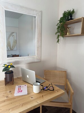 Ein Holztisch mit einem Laptop, einem Buch, einer Tasse und einem Brillenpaar. Dazu ein Stuhl mit Kissen, ein Blumentopf und ein gerahmtes Bild an der Wand.