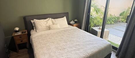 A bedroom with a large bed, wooden flooring, a nightstand, and a view through a glass door to an outdoor area.