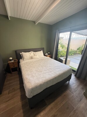 Ein Schlafzimmer mit einem großen Bett, Holzboden, einem Nachttisch und Blick durch eine Glastür in einen Außenbereich.