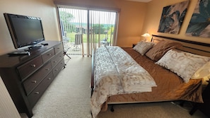 A bedroom with a bed, a television on a dresser, and a view of the outdoors through a sliding door. - Charming 2BR Condo in Lake Geneva - Walk to Pool, Tennis & Downtown! (Lake Geneva)