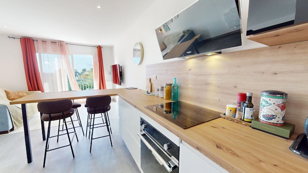 A modern kitchen with a wooden countertop, bar stools, a built-in oven, and a large window with curtains. - Le Cocon de la Paludiere (Le Pouliguen)