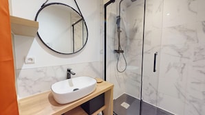 A modern bathroom with a white marble sink, a round mirror, and a glass-enclosed shower. - Le Cocon de la Paludiere (Le Pouliguen)