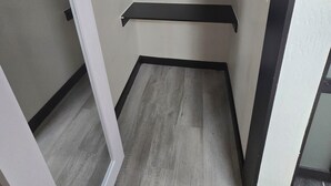 Ein schmaler, geschlossener Raum mit zwei schwarzen Regalen an der Wand, einem weißen Türrahmen und einem grauen Holzboden.