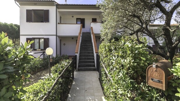 Un edificio de dos pisos con una escalera que conduce a la entrada, rodeado de vegetación.