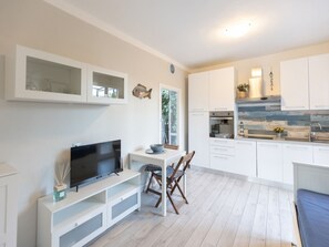 Eine moderne Küche mit weißen Schränken, ein Essbereich mit Fernseher und Sitzgelegenheiten sowie ein Blick ins Freie.
