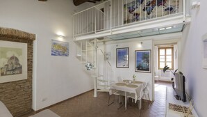 A spacious living area with a high ceiling, a staircase, a dining table with chairs, and a view of the outdoors through a window. - In the heart of Portoferraio's historic center! (Portoferraio)