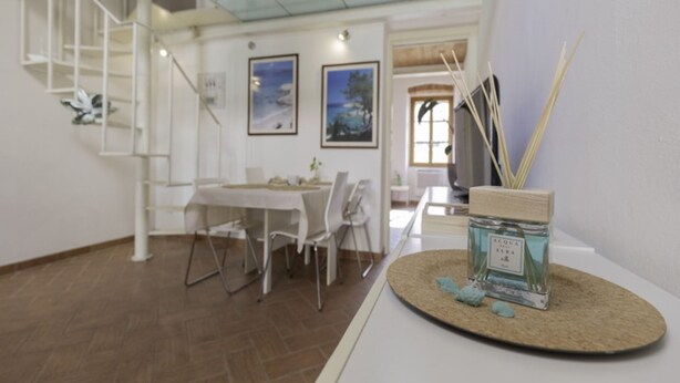 A spacious room with a high ceiling, a glass staircase, a dining area with white furniture, and a table with a decorative jar. - In the heart of Portoferraio's historic center! (Portoferraio)