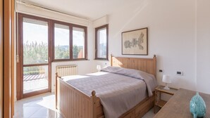 A bedroom with a wooden bed, a nightstand, a lamp, and a view of the outdoors through large windows. - Spacious, bright and comfortable with 2 terraces (Portoferraio)