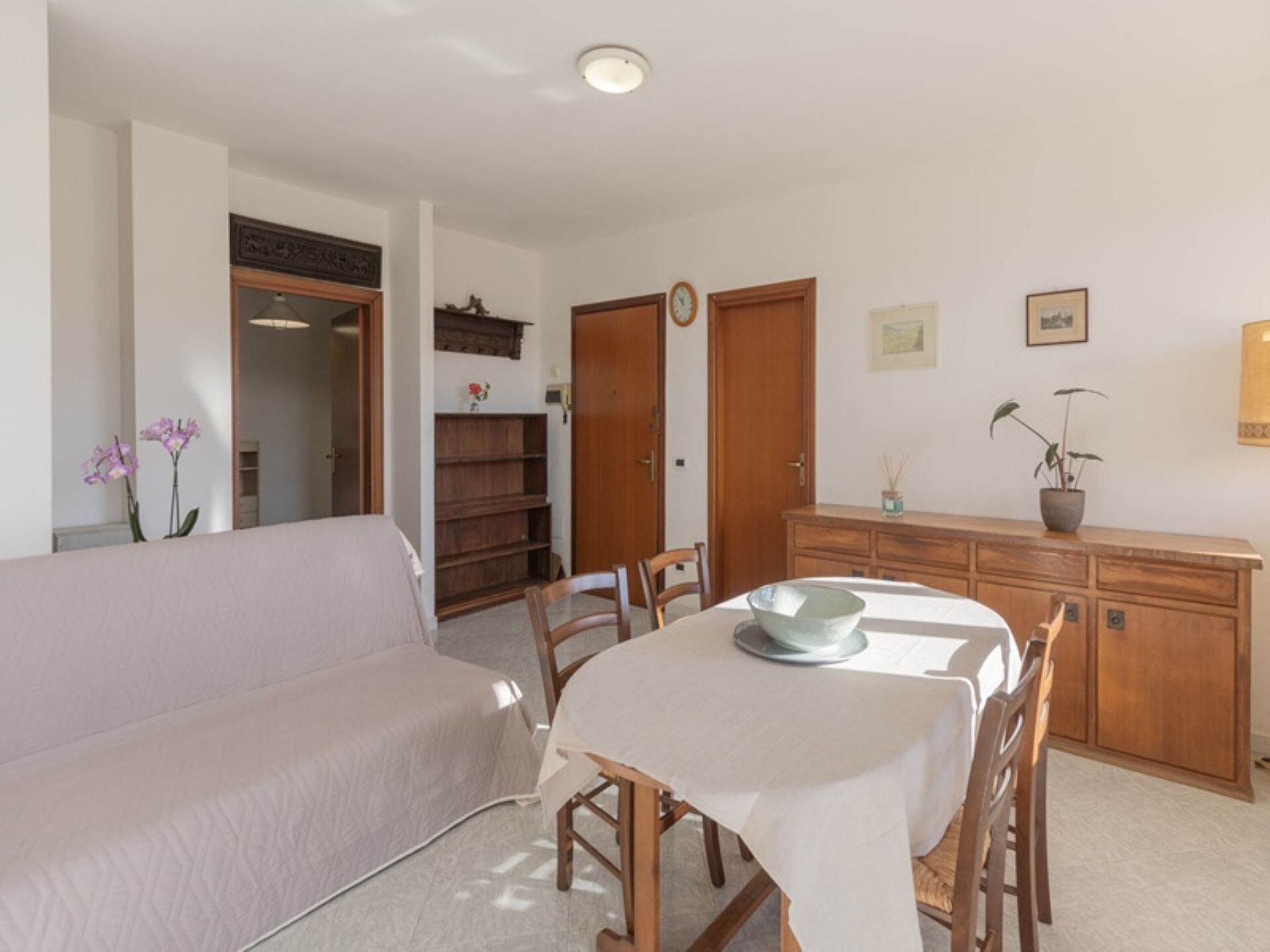 Un comedor con una mesa dispuesta para cuatro personas, rodeada de sillas de madera. A la izquierda hay un sofá y un armario de madera con un reloj y objetos decorativos en la pared detrás de la mesa.