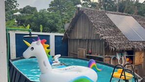 Een buitenzwembad met opblaasbare eenhoorn en regenboog drijvers, omgeven door een rieten hut en weelderige begroeiing.