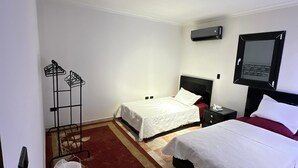 Habitación de hotel con dos camas, piso a cuadros rojo y beige, un cubo blanco, una mesita de noche negra y aire acondicionado instalado en la pared.