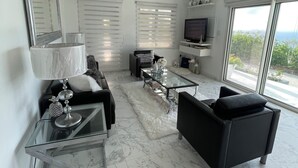 A modern living room with a glass coffee table, black sofas, a TV, and a ceiling fan. - Modern Ocean View Apartment (Pointe Blanche)