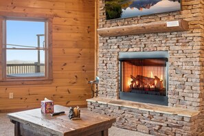 Ein Kamin aus Stein mit klarem Glasschirm, ein Holztisch mit einer Kiste und einer Fernbedienung sowie ein Fenster mit Blick auf eine Holzterrasse.