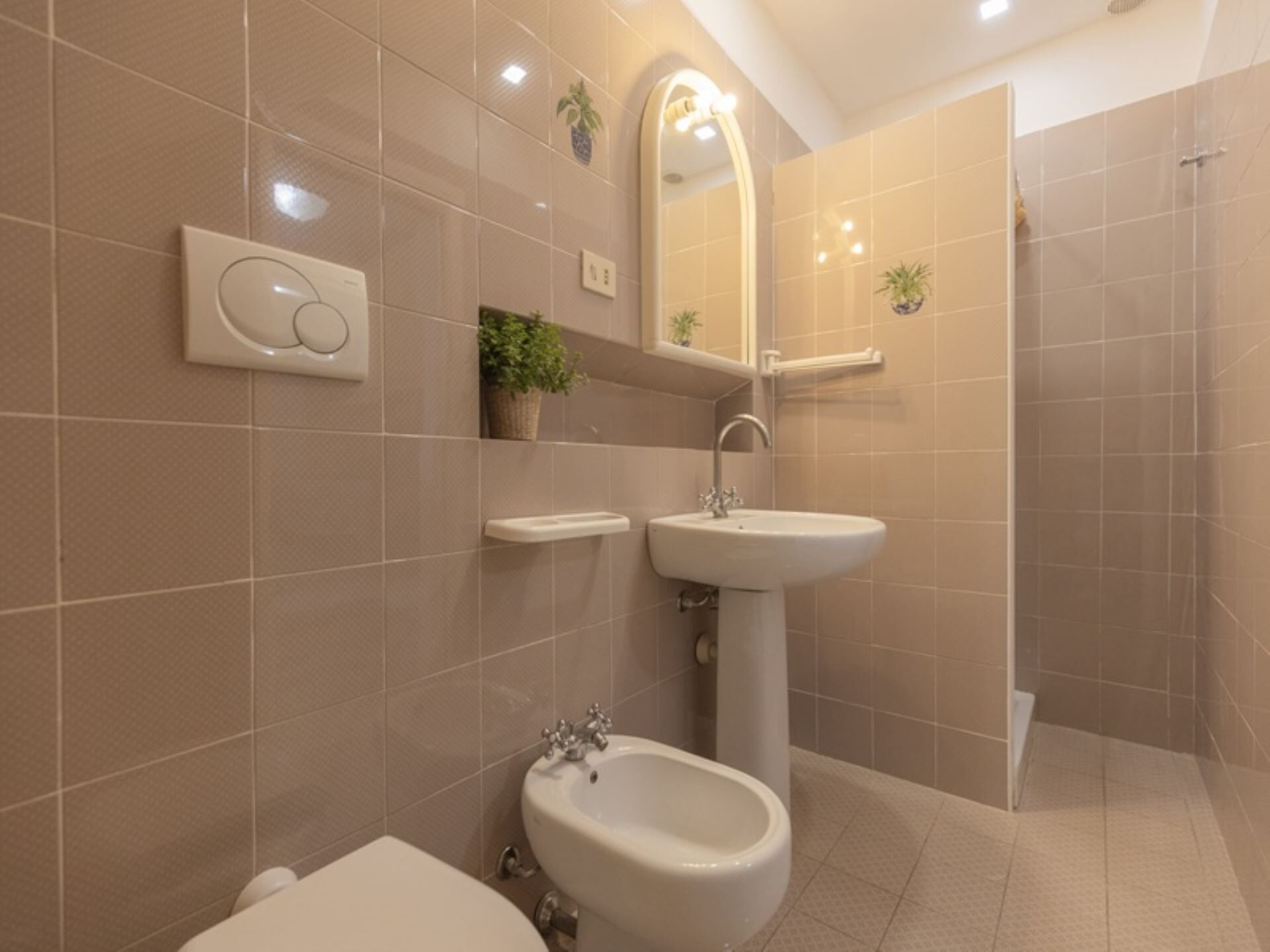 A modern bathroom with a pedestal sink, a bidet, and a toilet. There are potted plants on the wall and a mirror above the sink.
