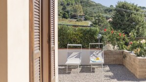 A balcony with two white lounge chairs, a stone-tiled floor, and a view of a hillside with greenery and flowers. - One-bedroom apartment with garden & Lacona view (Lacona)