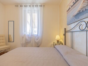 Un dormitorio con cama, una mesita de noche con lámpara, una silla, un espejo y una ventana con cortinas ligeras.