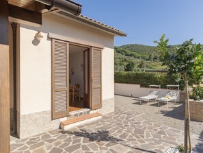 Una casa con patio de piedra, contraventanas de madera y vistas a una ladera verde.