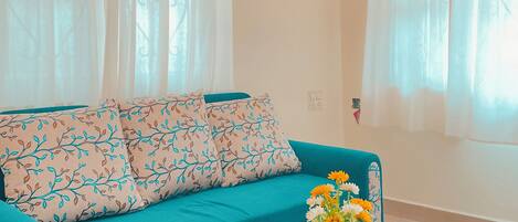 En teal farvet sofa med mønstrede puder, et glas sofabord med en fjernbetjening og en vase med blomster.