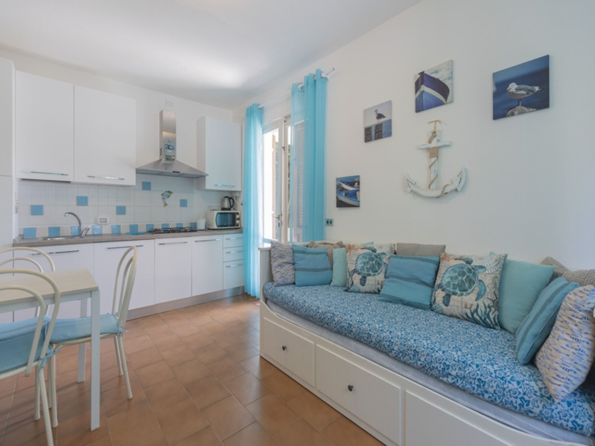A compact kitchen with white cabinets, a dining table with chairs, and a sofa with nautical-themed cushions.