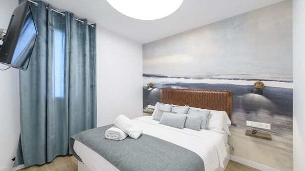 A neatly made bed with a grey blanket and white pillows, a wall-mounted TV, and a large window with blue curtains. - GRAN VIA capitals, LOS ANGElES (Madrid)