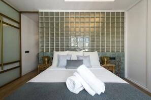 A neatly made bed with white linens, a grey throw, and a folded towel. The room has a large window with a view, a wooden floor, and a wall with a grid pattern. - GRAN VIA (all caps), MaRRAKECH (Madrid)