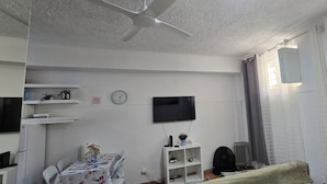 A living room with a ceiling fan, a flat-screen TV mounted on the wall, a clock, and a radiator. - Basement apartment studio in Lisbon Portugal, near the center. (Lisboa)