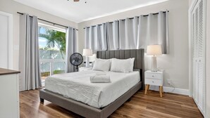 A bedroom with a large bed, two bedside lamps, a ceiling fan, and a view of the outdoors. - Swaying Palms Unit 1: 2 BR 2 Bath Retreat, Near Beach, Heated Pool, Free Parking (Fort Lauderdale)