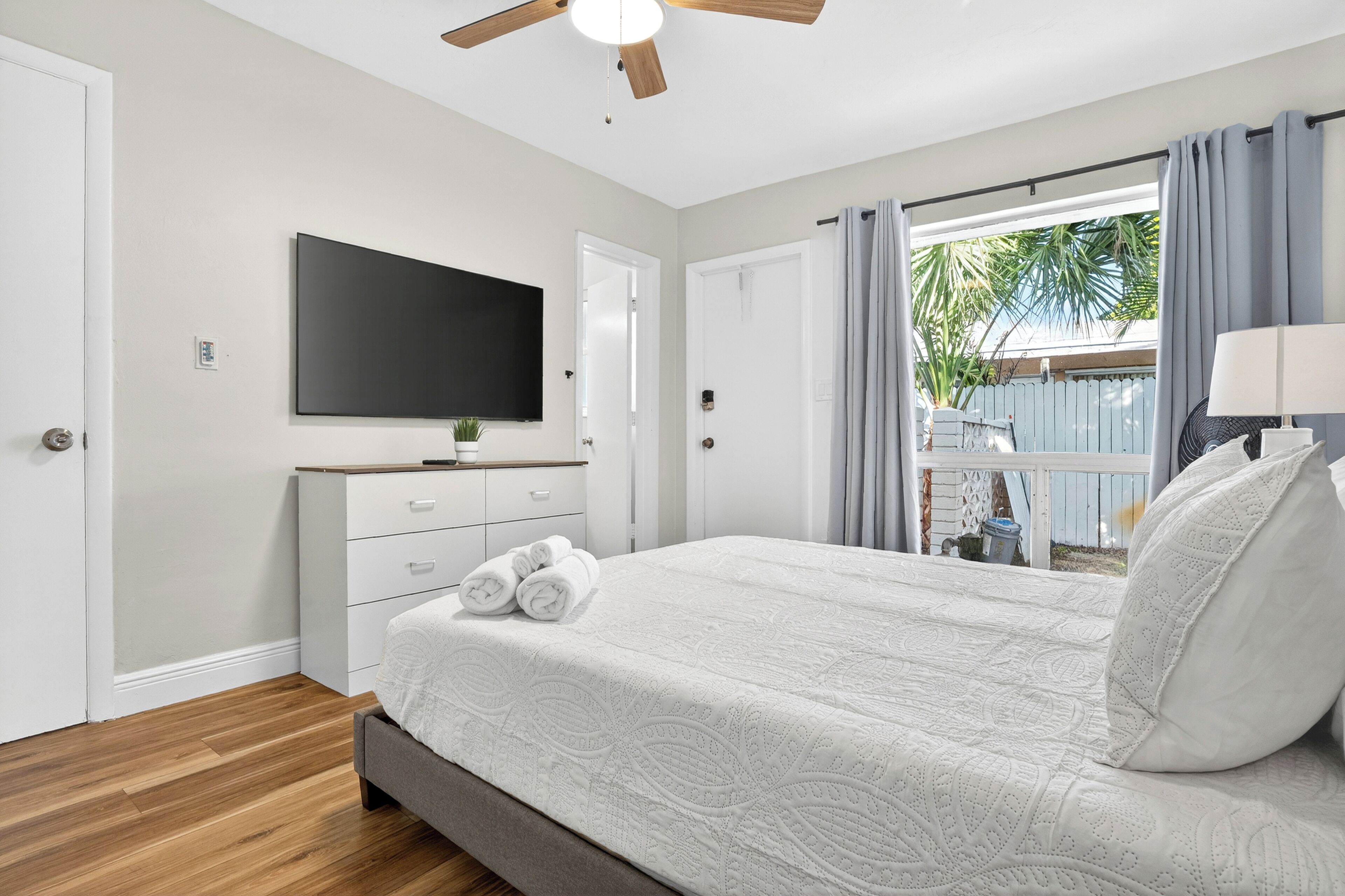 Un dormitorio con cama, ventilador de techo, televisión y ventana con cortinas.