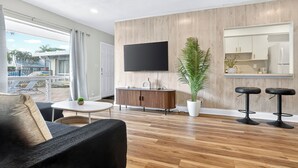 A modern living room with a flat-screen TV, wooden panel walls, a potted plant, a dining table, and two bar stools. - Swaying Palms Unit 1: 2 BR 2 Bath Retreat, Near Beach, Heated Pool, Free Parking (Fort Lauderdale)